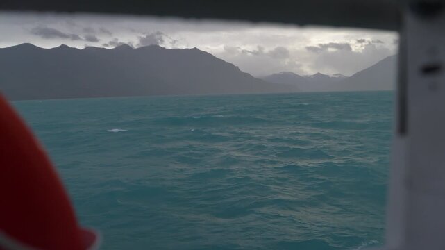 Lago Argentino, Santa Cruz, Argentina, featuring rough turquoise waters, strong winds, overcast skies and rugged Patagonian Andes during a lake crossing within Los Glaciares National Park onboard shot