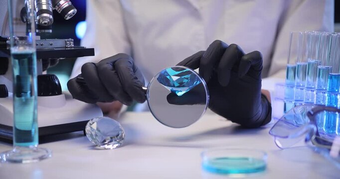 Scientist inspects blue diamond through magnifying glass in laboratory. Black gloved hands angle gemstone studying cut clarity with steady precision