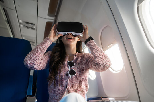 Young woman wearing a virtual reality headset while sitting by an airplane window, pointing outside during a flight. Modern travel, technology and future experience concept.
