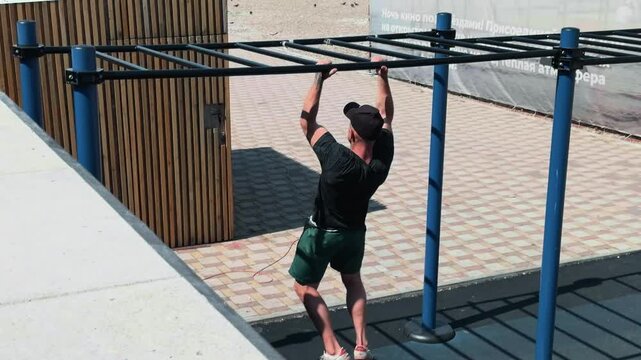 Fit Caucasian athlete in black tank top and backward cap doing pull-ups on blue bar of outdoor calisthenics gym rig in sunny resort park. Medium shot authentic effort and back muscles