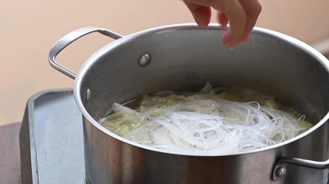 Woman hand putting glass noodles into boiling pot of Chinese cabbage soup with pork, Asian food cooking