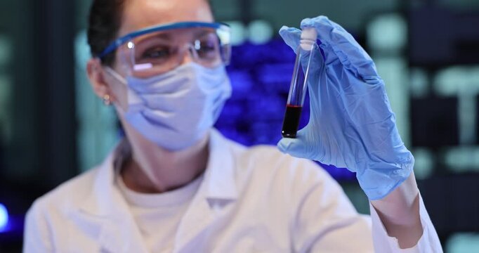 Woman scientist studies dark liquid inside glass tube in lab. Gloved hand lifts sample toward bright light to check color change carefully slow motion