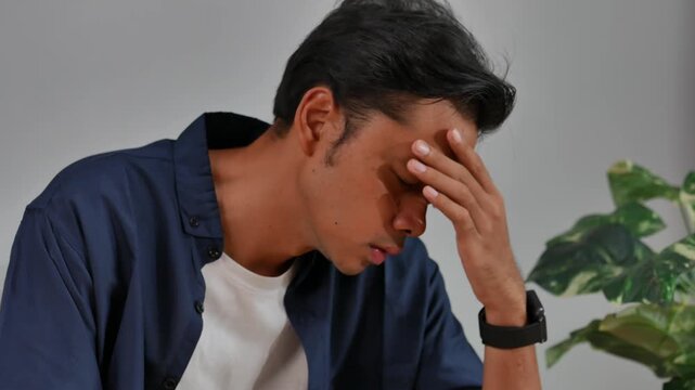 A young man sitting on the sofa in the living room of his house, with a sad, confused, dizzy, stressed expression while massaging his head.