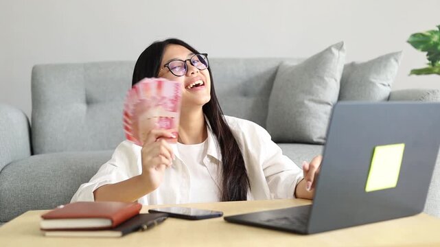 A beautiful smiling Asian woman sits on the floor of her living room with her laptop on the table while holding a stack of cash bills making a profit or income from her online business.