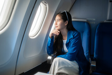 Fototapeta premium Young woman taking a selfie by the airplane window during a flight, smiling in a blue jacket while seated in a passenger seat, showing travel, leisure, and modern lifestyle