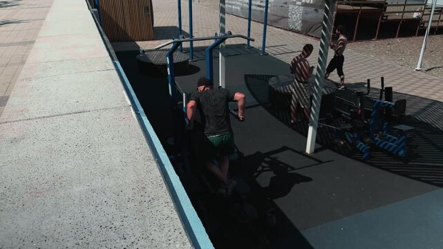 High angle view of athletic man doing dips on parallel bars at outdoor street workout park. Concrete parapet in foreground creates perspective. Slatted canopy casts geometric striped shadows