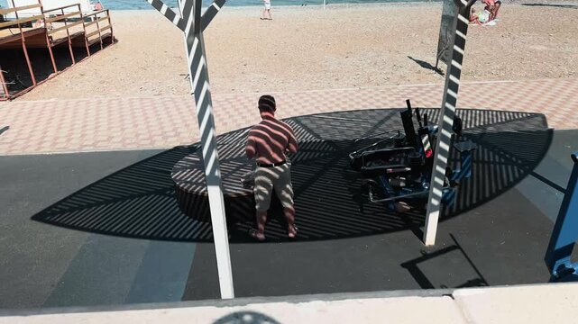 Unrecognizable shirtless man standing by circular bench at outdoor beach gym under slatted canopy. Striking geometric striped shadows pattern on his bare back. Handheld tracking wide observer shot.