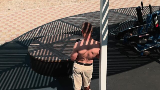 Unrecognizable shirtless muscular man standing at outdoor beach gym leaning on white pillar. Athlete looking at smartphone with striking geometric striped shadows on bare back during hot summer day