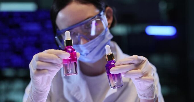 Woman dermatologist compares pink cosmetic bottles in laboratory. Gloved hands tilt glass droppers checking liquid color during testing slow motion