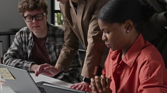 Multiethnic young programmers collaborating on laptop in office while manager gives directions. Ideal for tech startup marketing, software development, and corporate training videos