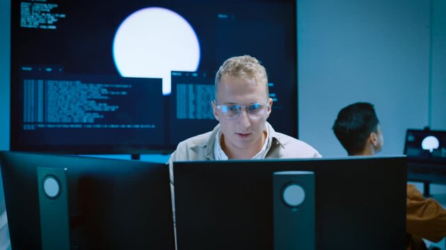 Focused young male IT specialist reviewing code and looking at computer monitors while developing AI software system in tech office