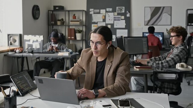 Young Asian programmer wearing glasses typing on laptop and drinking coffee in busy open space office. Footage for tech startup promotions and software development corporate videos