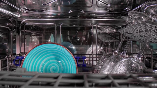 Camera pulls back from inside dishwasher revealing clean blue plates and wine glasses in rack.