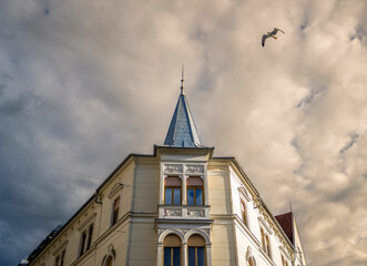 Obraz premium Historic corner building with soaring spire and flying bird against textured clouds creating timeless European street photography composition