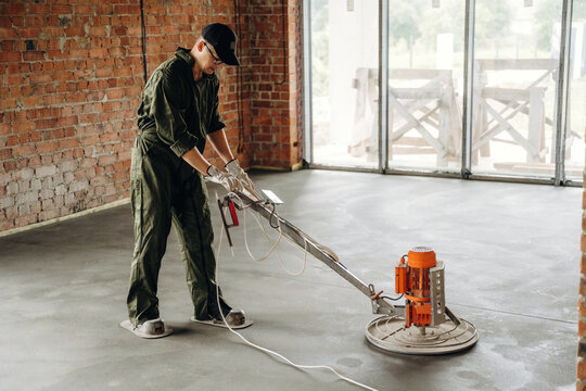 Male construction worker in green coveralls operates floor polishing machine on newly poured concrete in industrial space with exposed brick walls and large windows