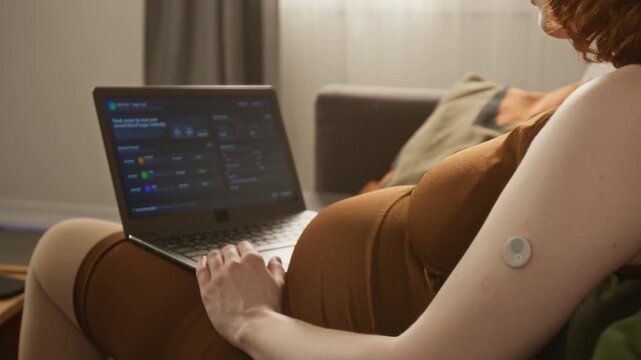Rack focus shot of pregnant woman tracking blood sugar levels on laptop, wearing continuous glucose monitor sensor on arm. Video perfect for gestational diabetes awareness and healthcare app marketing