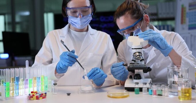 Woman chemist fills flask with powder as colleague looks in microscope at lab workplace. Female scientists in masks study pharmaceutical products