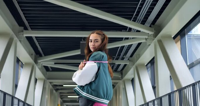 Young woman walking with earbuds and phone in modern corridor, holding cable and nodding to beat, varsity jacket and bright pants add stylish contrast against architectural beams, upbeat urban vibe