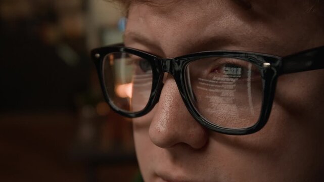 Extreme close-up shot of young male programmer wearing glasses focusing on screen with computer code reflection. Perfect for technology startups, cybersecurity ads, and software development promos