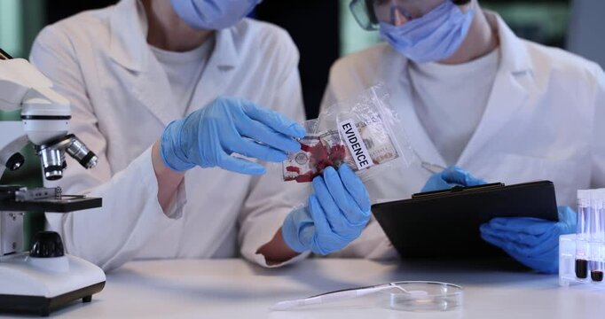Women experts study dollar bill with red stains in plastic bag at desk. Female forensic scientists document crime evidence investigation in lab