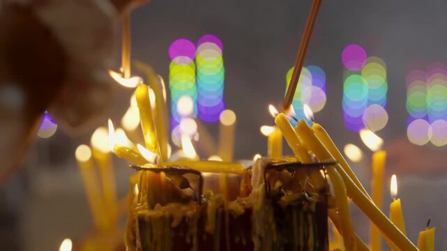 People are trying to light incense sticks from candles on a platform filled with candles behind a backdrop of colorful bokeh lights during the candlelit procession.