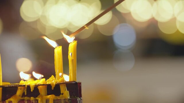 Incense sticks are lit next to a group of candles on a platform. In the background, there's a beautiful bokeh effect of lights. People are walking around during the candlelit procession.