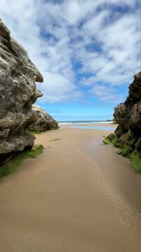 POV walk between coastal rocks across wet sand exposed by low tide toward an open beach under clear daylight.