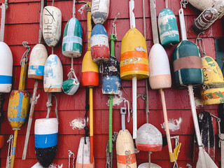 Collection of colorful, weathered lobster buoys hanging on a bright red fishing shack wall. Coastal maritime decor and New England fishing heritage captured in a detailed close-up. © Ryan