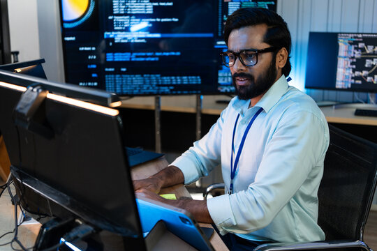 Indian software developer programming code on multiple monitor screens, managing corporate network security database and analyzing digital information at night