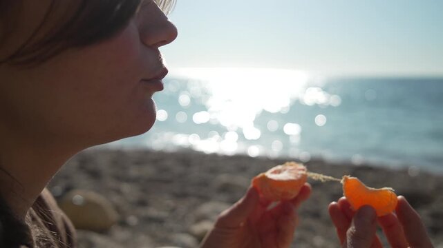 Eating tangerine beach woman enjoys a fresh mandarin slice while sitting on the sunny seashore during a peaceful summer vacation
