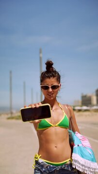 Young woman walking along a coastal road with smartphone and beach towel