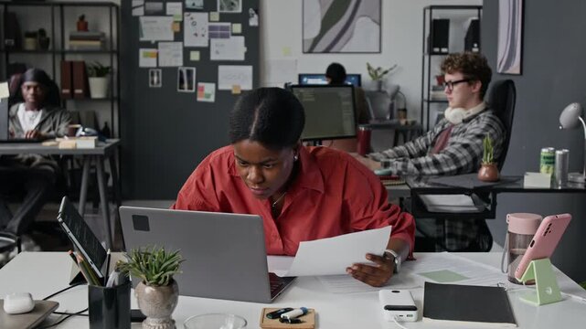 Young African American woman programmer feeling stressed, crumpling paper, closing laptop, holding head in hands in office. Video represents workplace burnout and software development errors