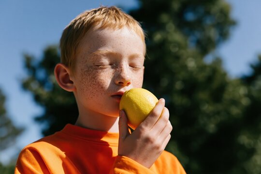Freckled child portrait with eyes closed holding a lemon against leafy garden backdrop in warm sunlight for natural health background and banner design