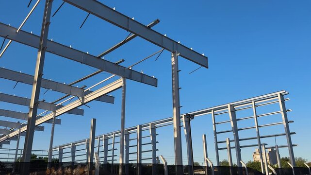 Steel frame building under construction on an open industrial site, with structural beams and columns against a clear blue sky, handheld pan