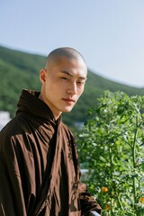 Obraz premium Standing shaved head man in brown robe beside balcony plants with hillside view for mindful lifestyle background and wellness banner