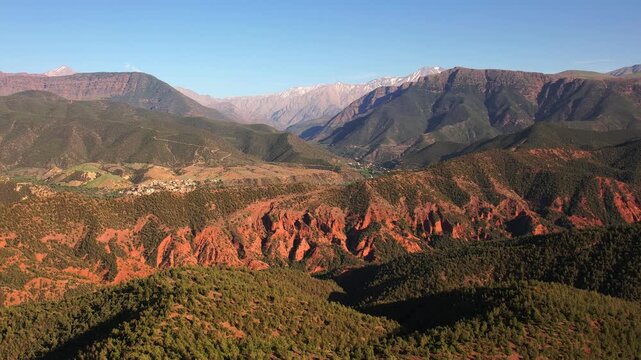 Ourika Valley, Al Haouz Province, Marrakesh Safi, Morocco, featuring red sedimentary badlands, juniper woodland, semi arid Mediterranean biome and snow covered High Atlas Mountains, drone zoom out