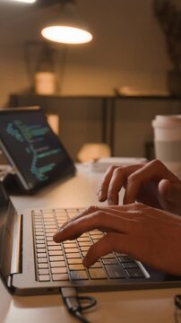 Vertical close-up video of anonymous programmer typing code on laptop keyboard with tablet displaying software scripts in background. Ideal for tech education or software development marketing
