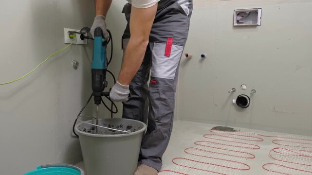 Construction worker preparing tile surface, mixing mortar substrate for flooring, underfloor heating mat. Workman combines adhesive in renovation setting. Operating power tool during bathroom upgrade