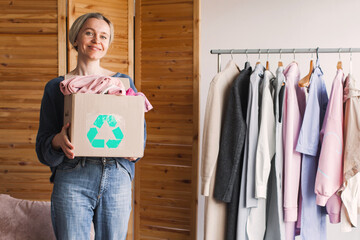Obraz premium Woman standing with a cardboard box full of clothes for recycling. Sustainability concept for garment donation. Eco friendly lifestyle choices, mindful consumption and fashion waste reduction.