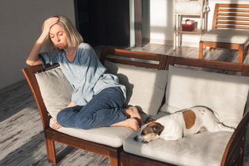 Obraz premium Sad woman sitting on couch with dog at home. Depressed female holding head while feeling exhausted. Loneliness and mental health struggle in domestic environment. Solitude and stress concept.
