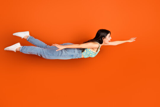 Cheerful woman in stylish casual outfit playfully poses as if flying against a vivid orange background, expressing happiness