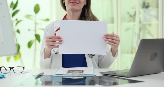 Woman doctor holds blank paper at clinic desk. Specialist presents empty sheet toward camera preparing space for clear medical message slow motion