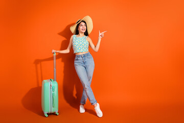Obraz na płótnie Canvas Young stylish female traveler posed against a vibrant orange background with suitcase ready for an exciting vacation