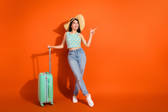 Young stylish female traveler posed against a vibrant orange background with suitcase ready for an exciting vacation