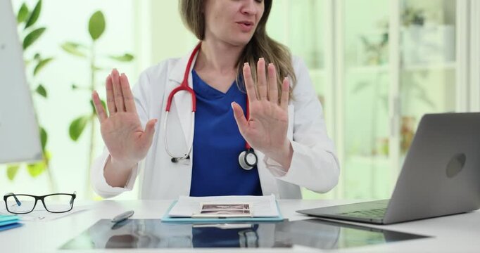 Woman doctor raises hands in No gesture at clinic desk. Specialist moves palms forward refusing request during calm medical appointment slow motion