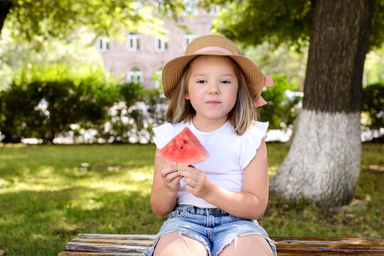 Little girl in a straw hat holding a watermelon slice while sitting on a park bench. Healthy summer snack, childhood happiness, outdoor lifestyle, and seasonal family recreation