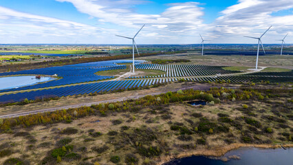 Wide aerial drone view of a modern solar farm with rows of blue photovoltaic panels and tall wind turbines scattered across green fields under a blue sky with distinctive clouds. © Adam