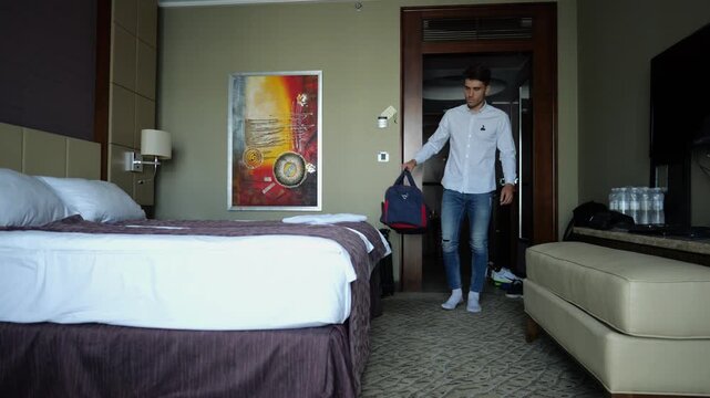 Tired young businessman entering modern hotel room with travel bag and falling onto bed after long business trip. Exhausted man relaxing in comfortable accommodation interior after work and travel
