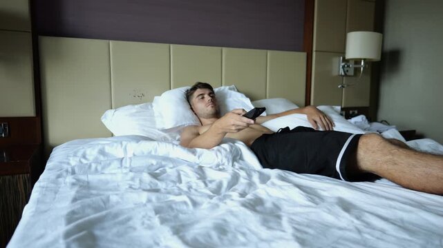 Young shirtless man switch channels on TV remote control lying on large bed in modern hotel room or home interior. Relaxed guy enjoying leisure, entertainment and comfortable lifestyle indoor