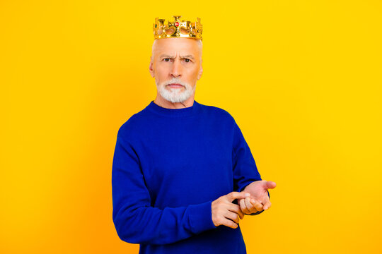 Elderly man wearing a gold crown and blue sweater posing serious and regal against a bright yellow background, portrait of a proud kinglike character with confident expression